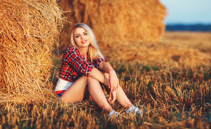 Блондинка в красной рубашке сидит возле стога сена
